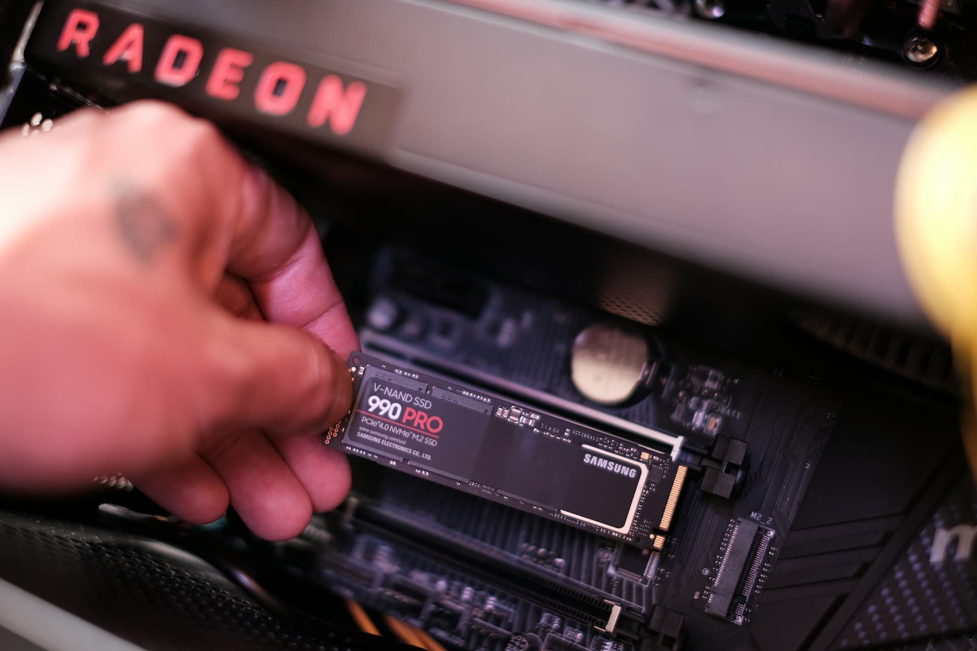 Hand inserting a Samsung 990 Pro M.2 NVMe SSD into a motherboard slot with AMD Radeon GPU visible