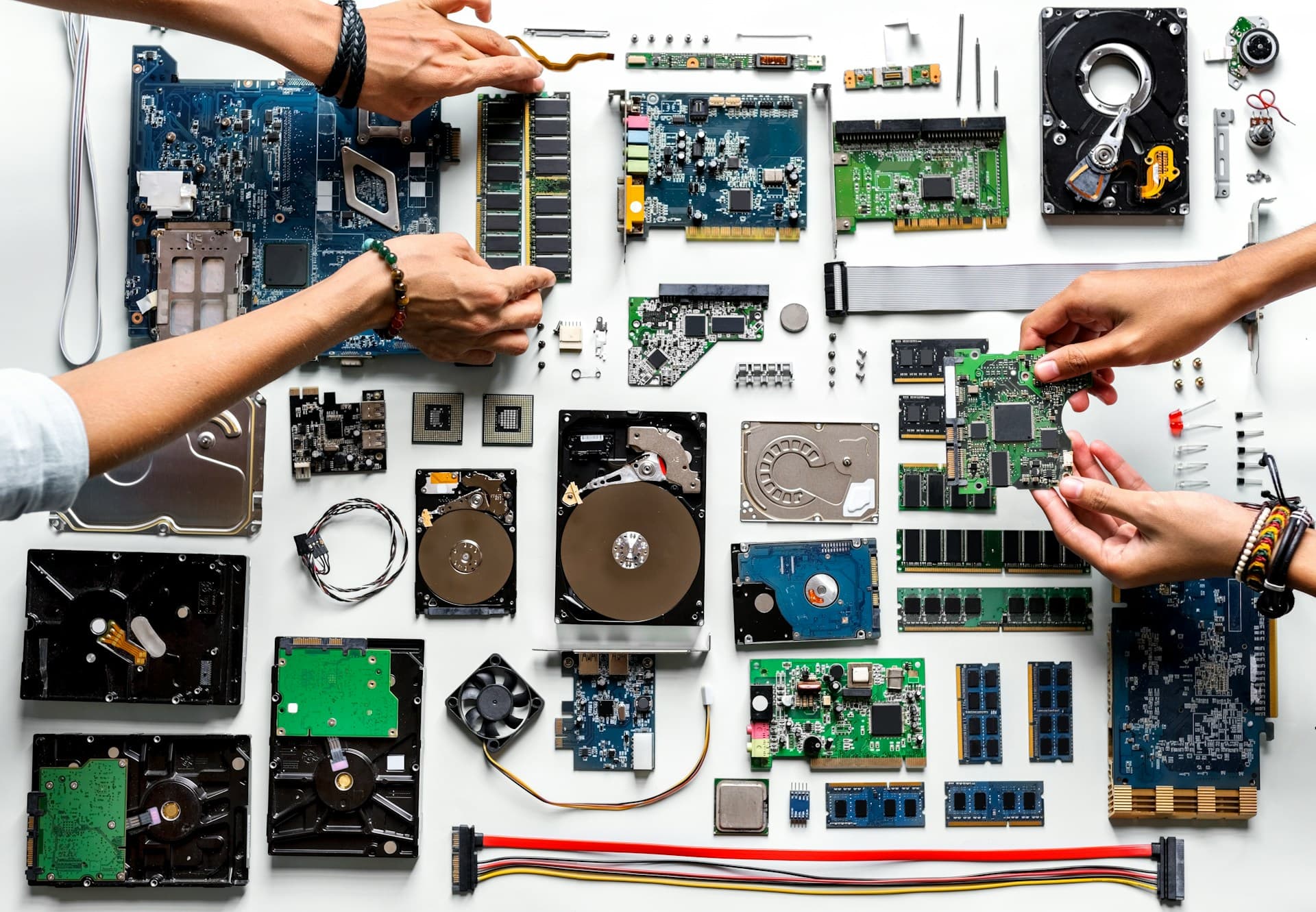 Overhead view of various old PC components including hard drives, RAM, motherboard and expansion cards laid out on a white surface