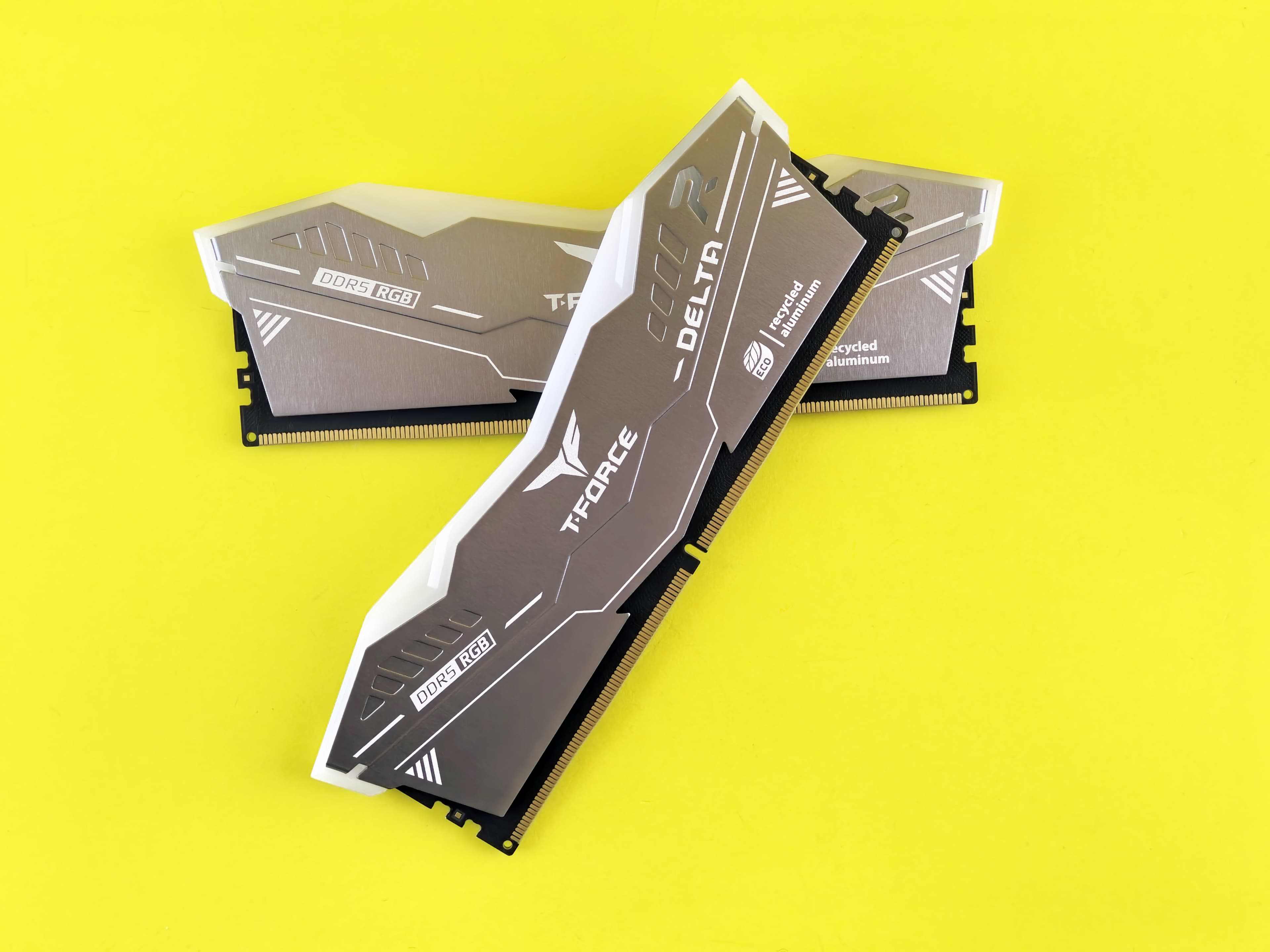 Two TeamGroup T-Force Delta DDR5 RGB 32GB RAM sticks with silver brushed aluminum heatspreaders, crossed over each other on a bright yellow background, featuring ECO recycled aluminum branding and gold edge connectors.