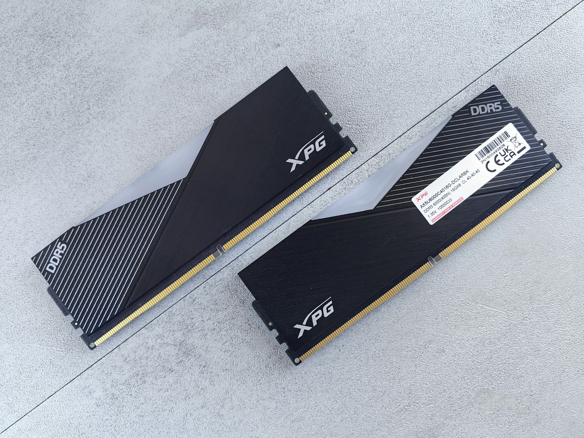 Two ADATA XPG DDR5 6000MHz RAM sticks with black heatspreaders laid flat on a grey surface, showing DDR5 branding and gold edge connector