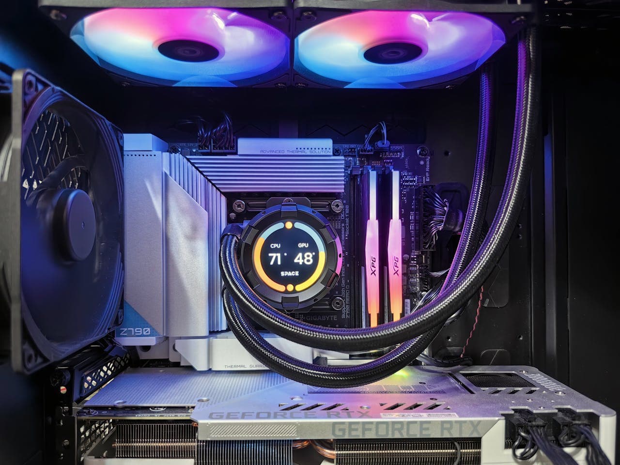 Inside view of a PC case with side panel removed, showing a Gigabyte Z790 motherboard with two XPG DDR5 RAM sticks installed, an NVIDIA GeForce RTX GPU in the PCIe slot, an AIO liquid cooler displaying CPU and GPU temperatures, and RGB fans illuminated in blue and purple